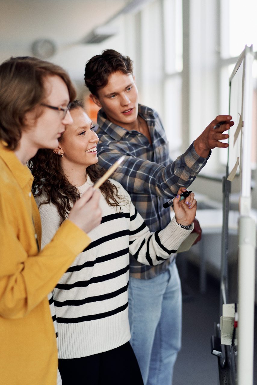 Tre studenter står och planerar vid en whiteboardtavla