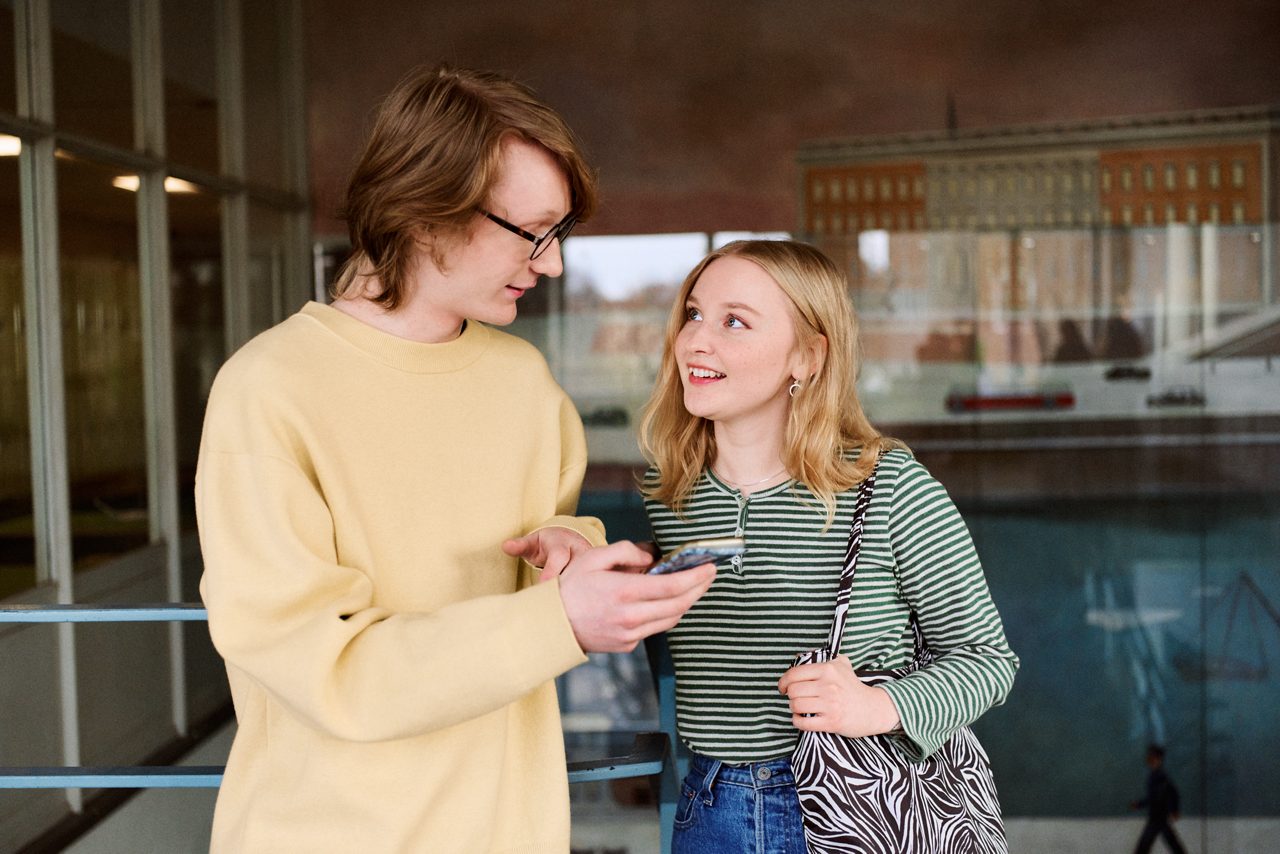 Två studenter står och pratar och tittar på en mobiltelefon