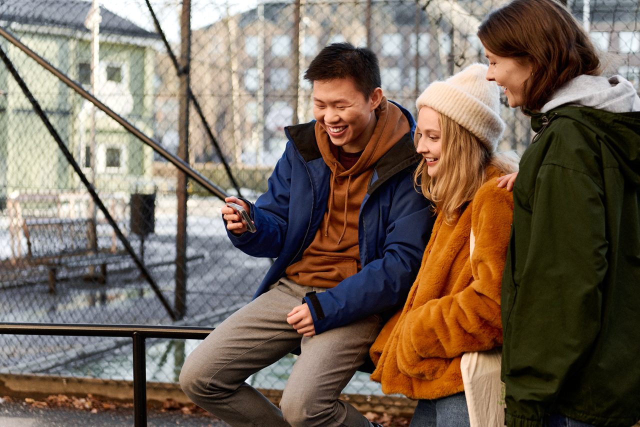 Tre studenter hänger på skolgården, tittar på en mobiltelefon och skrattar