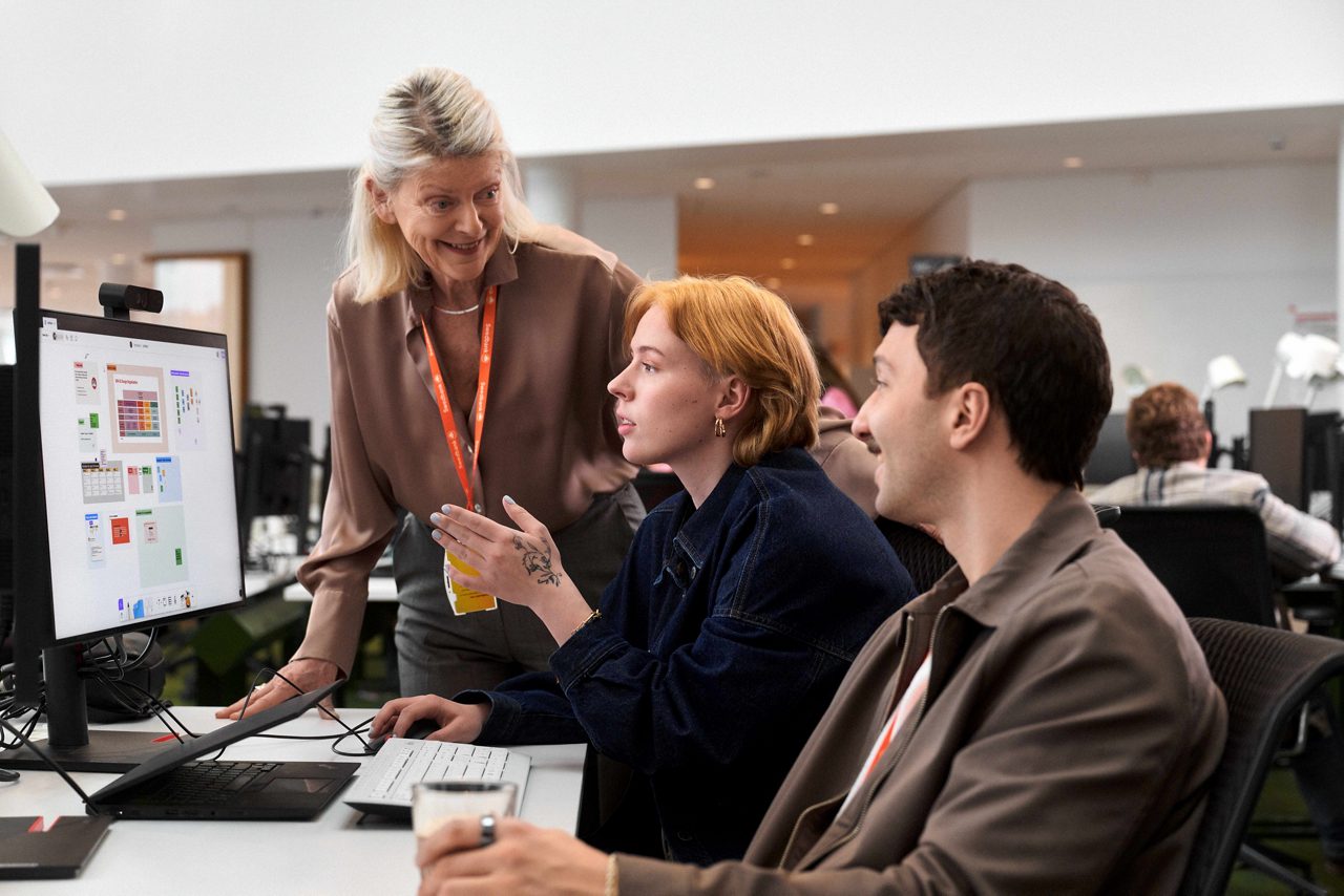 Two younger people sitting infront of a computer, talking to a person standing beside the desk