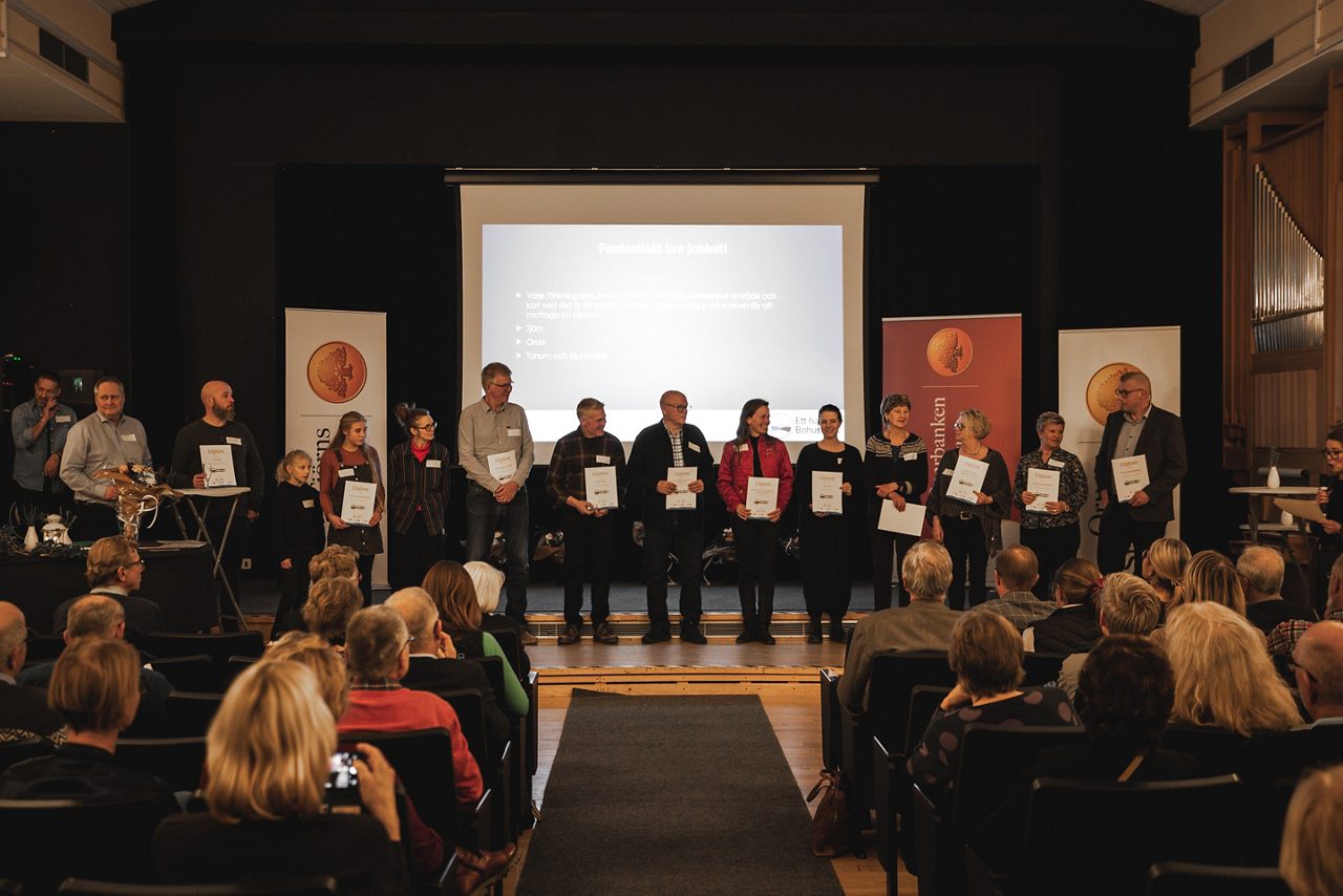 En grupp personer står på scen inför publik och håller diplom vid ett evenemang arrangerat av Orusts Sparbank.