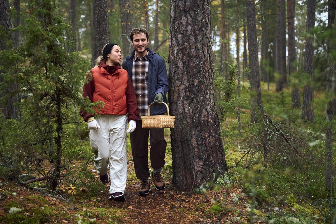 A woman and a man walking along a forest path, one carrying a woven basket. 