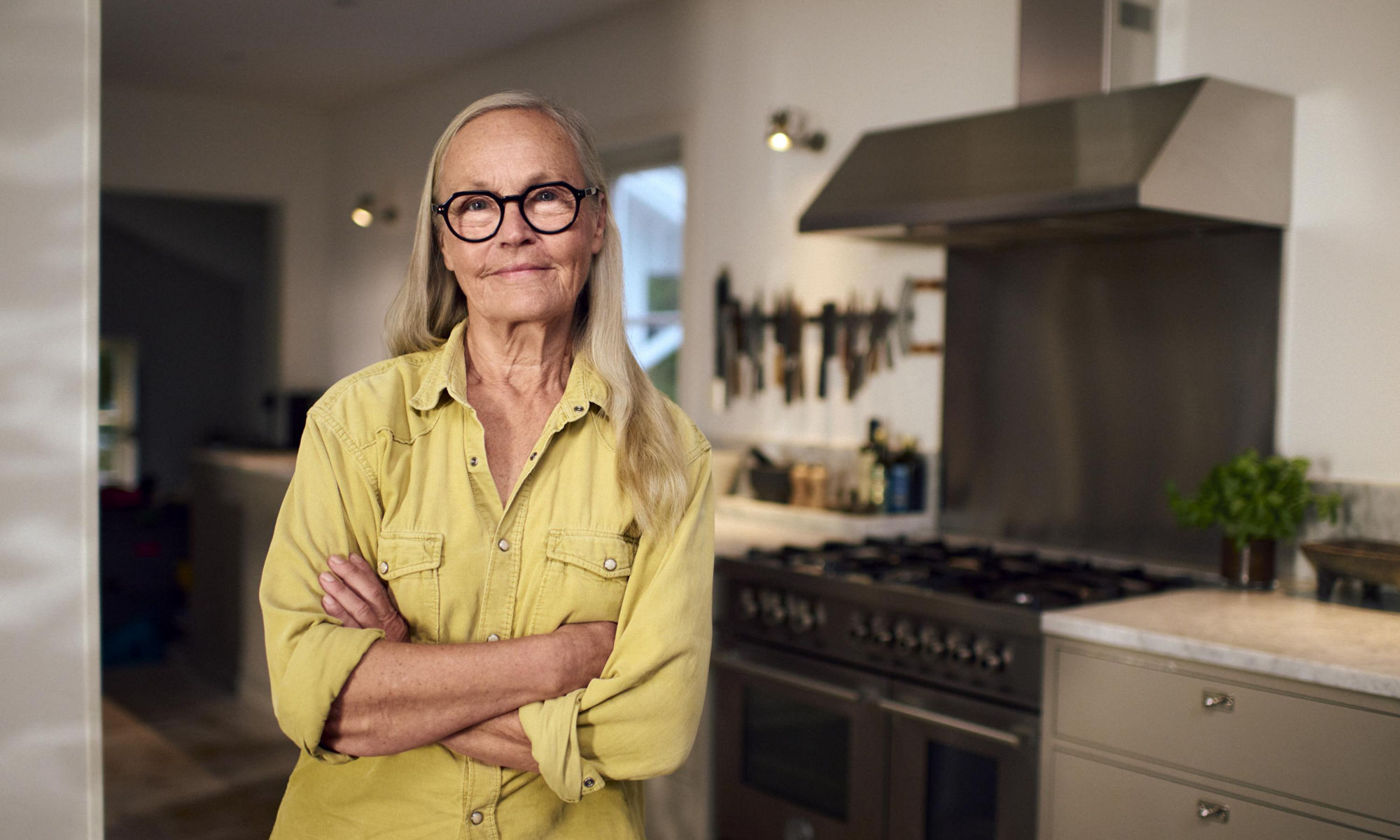 En självsäker äldre kvinna står med armarna i kors i ett kök.