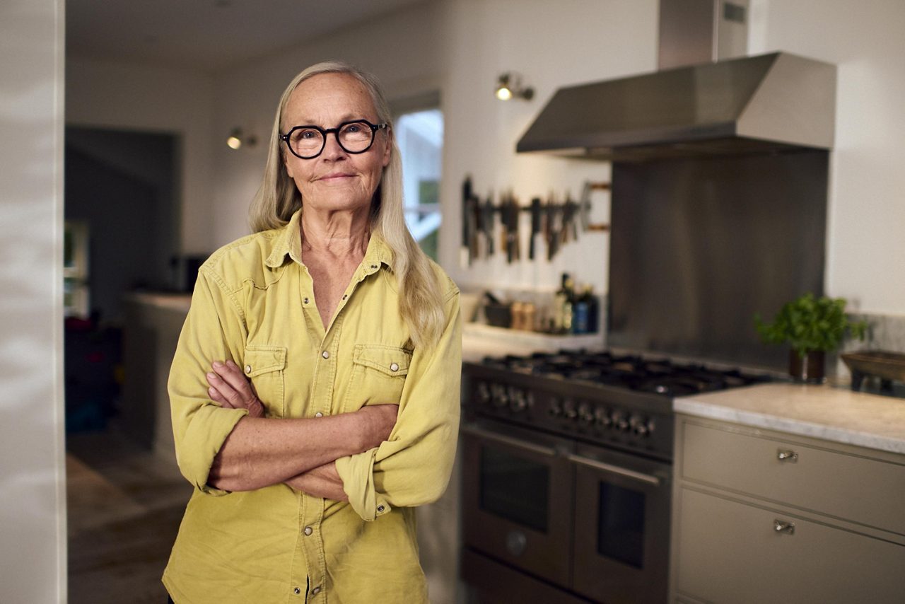 En självsäker äldre kvinna står med armarna i kors i ett kök.