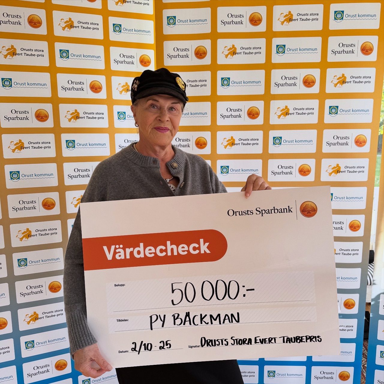 Py Bäckman håller en stor symbolisk check på 50 000 kronor framför en fotovägg med Orusts Sparbank och Orusts kommun.
