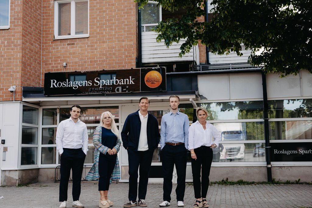Fem personer står framför entrén till Roslagens Sparbank. I bakgrunden syns bankens skylt, stora fönster och en tegelbyggnad.