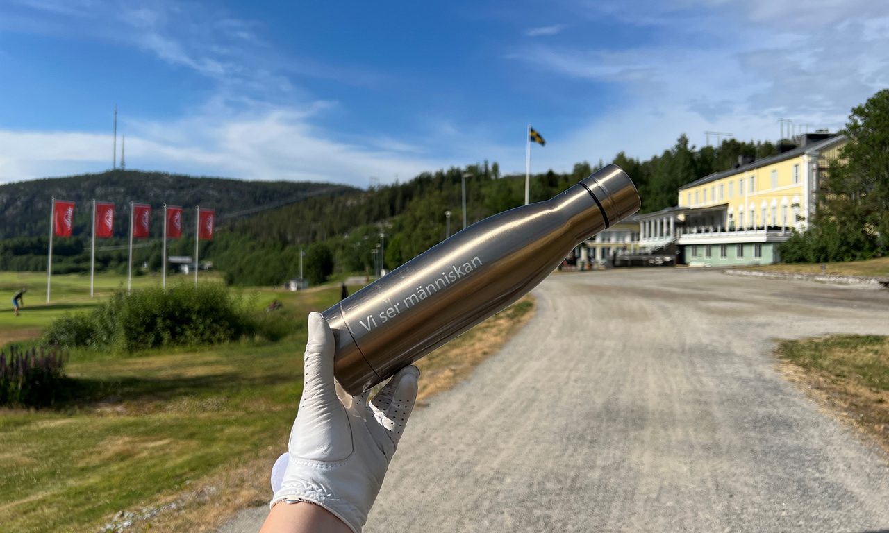 Hand med golfhandske håller upp en vattenflaska med texten "Vi ser människan"