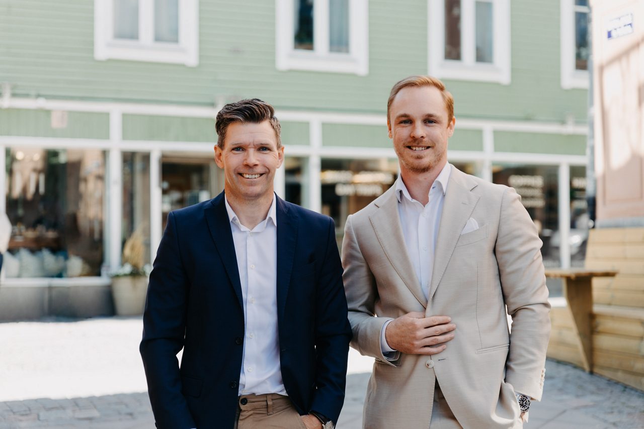 Två leende män i kavaj står utomhus framför en modern byggnad.