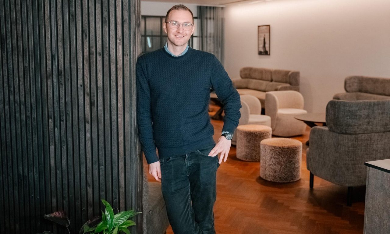 Leende man lutar mot vägg i modern lounge med fåtöljer och mjuk belysning.
