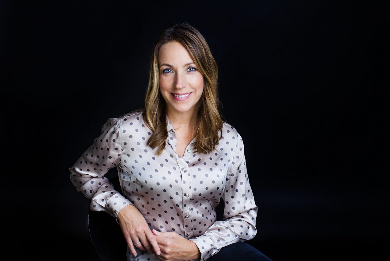 Bild på psykolog Anna Bennich. Porträttbild av en leende kvinna med långt ljusbrunt hår och blå ögon, klädd i en ljus prickig blus, sittande mot en mörk bakgrund i en professionell studiofotografering.