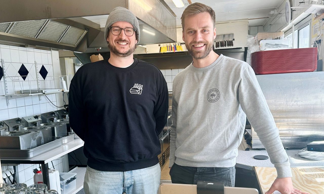 Två män står sida vid sida, leende mot kameran, i ett restaurang kök