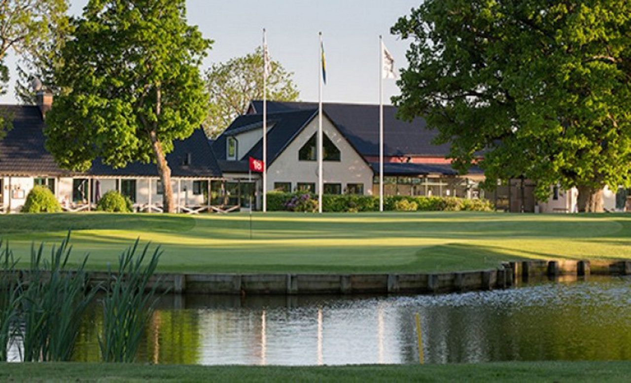 A well-maintained golf green sits beside a calm water hazard in front of a clubhouse building. 