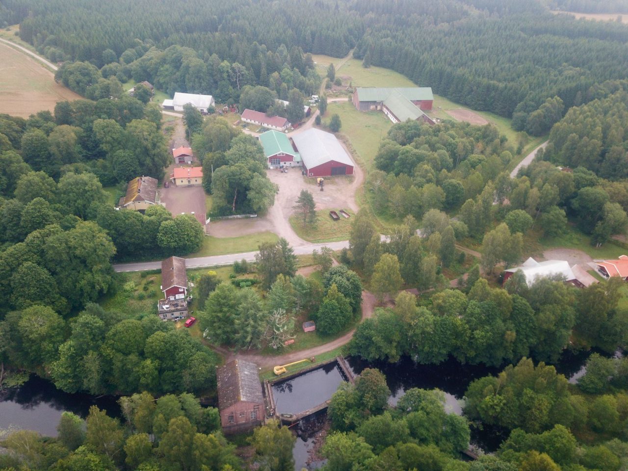 Flygfoto över gård där man ser hus, vatten och skog.