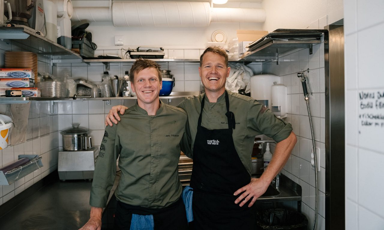 Två kockar står leende i restaurangkök med rostfria bänkar och hyllor.
