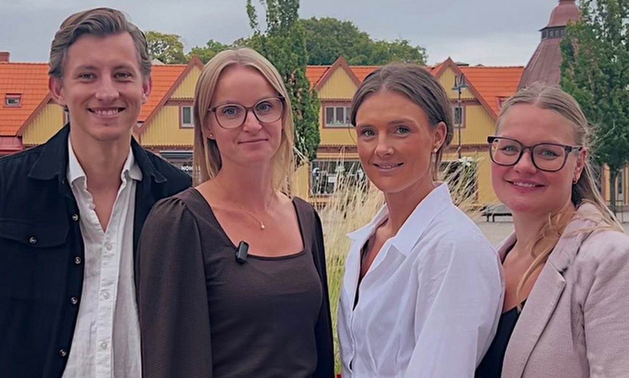 Fyra personer står på ett torg och tittar in i kameran.