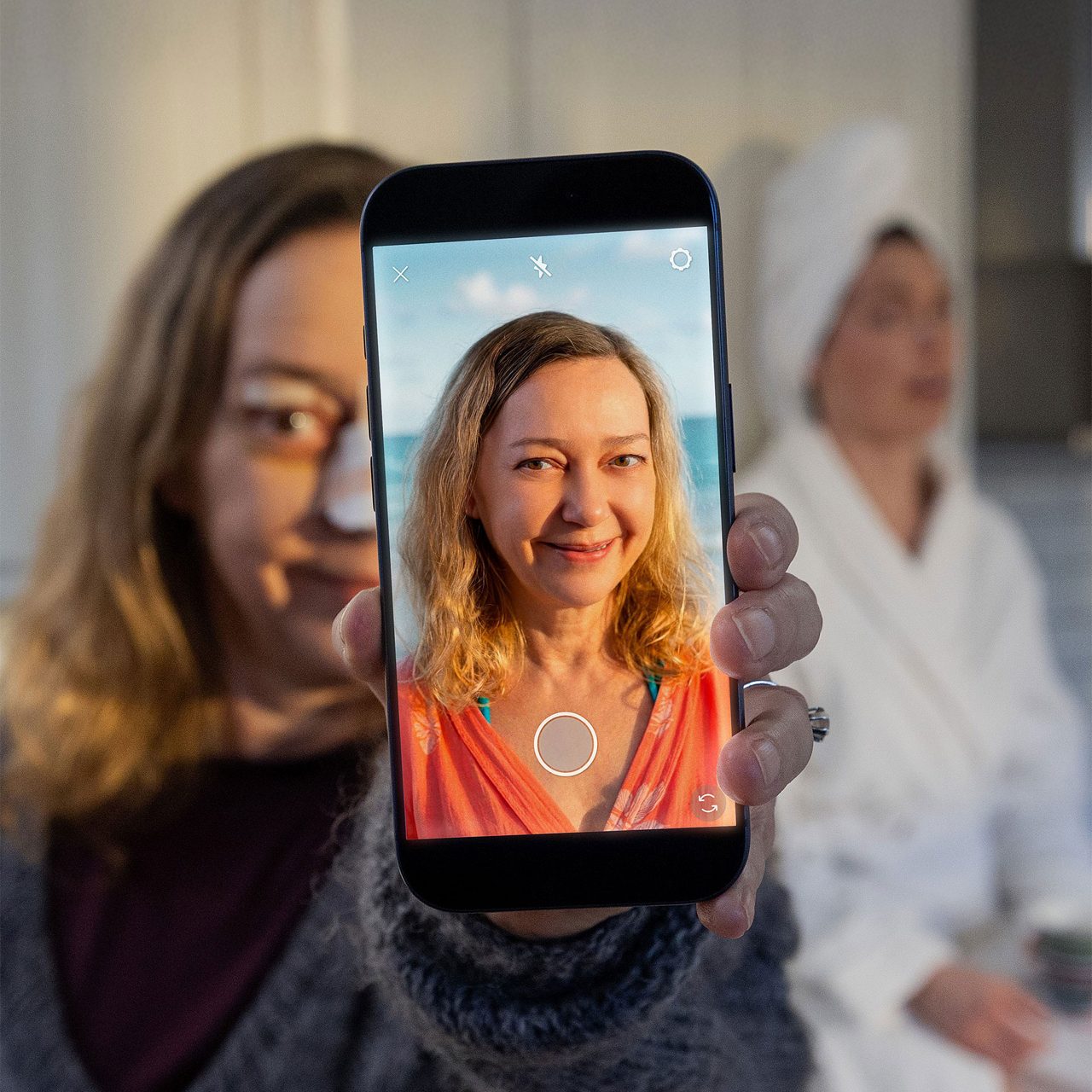 A woman holds a smartphone in front of her face, showing a bright, smiling selfie with a beach background on the screen. The real-life setting appears to be a home interior, with another woman in the background wearing a towel wrap, suggesting a casual or self-care moment. The contrast between the warm, sunny selfie and the cooler indoor lighting highlights the difference between online persona and reality. No visible text or numbers appear on the phone screen or in the environment.