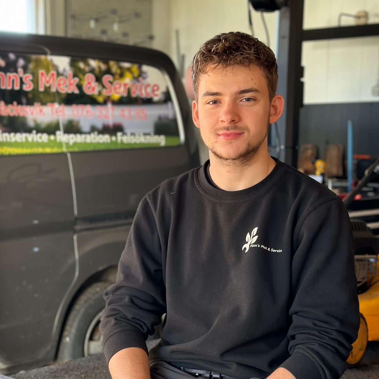 Finn sitter ner och tittar i kameran, i bakgrunden syns firmabil med text Finn's Mek och Service