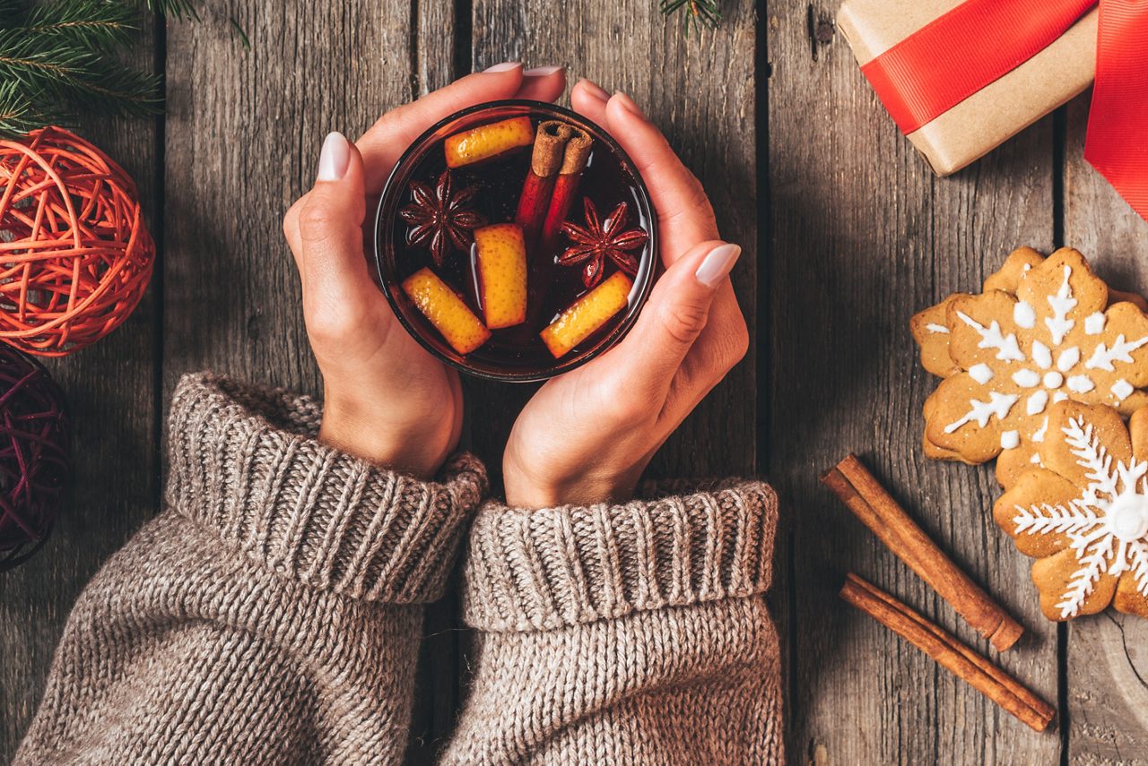 Två händer håller om en kopp varm juldryck, en julklapp och pepparkakor ligger på bordet