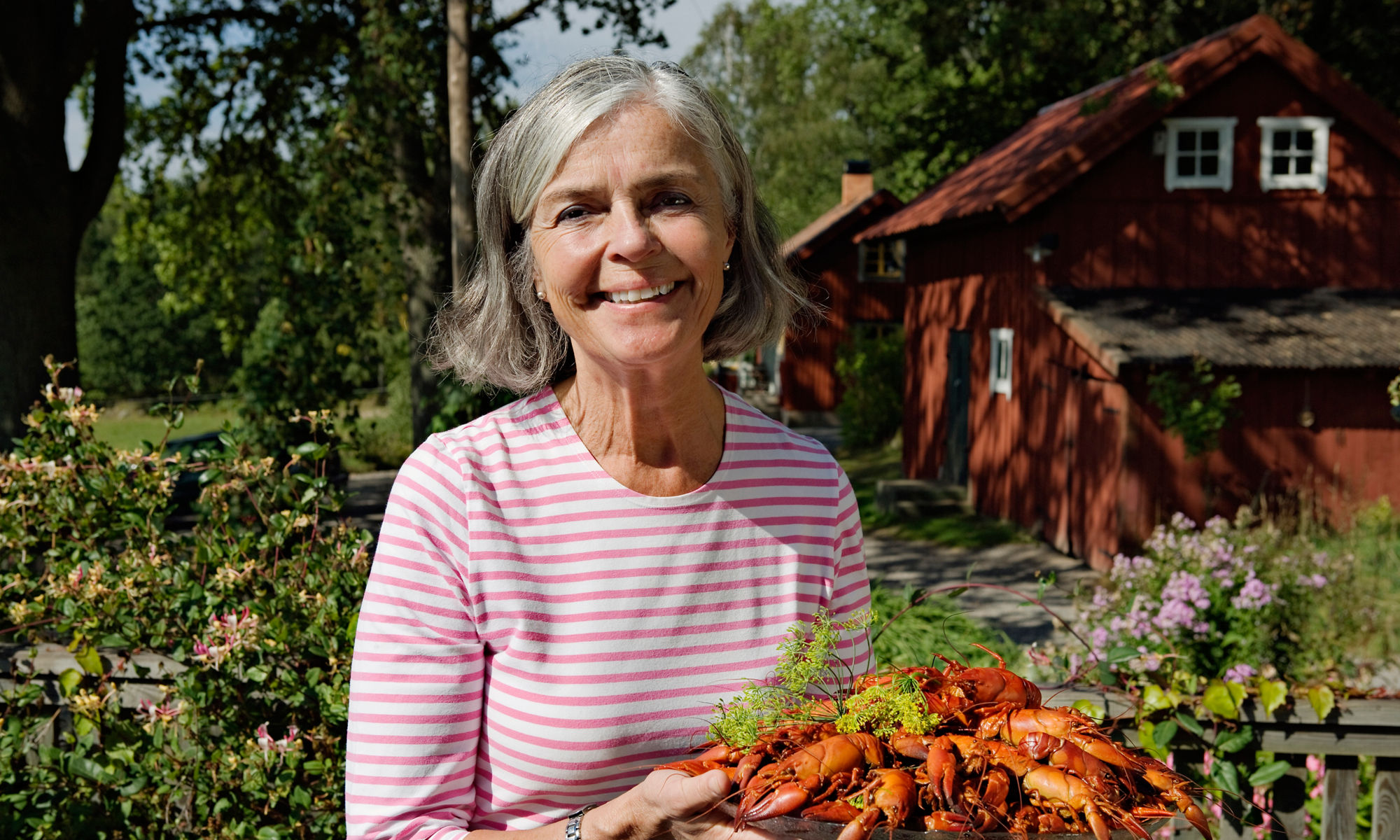 En äldre kvinna står i en trädgård och håller ett fat med kräftor