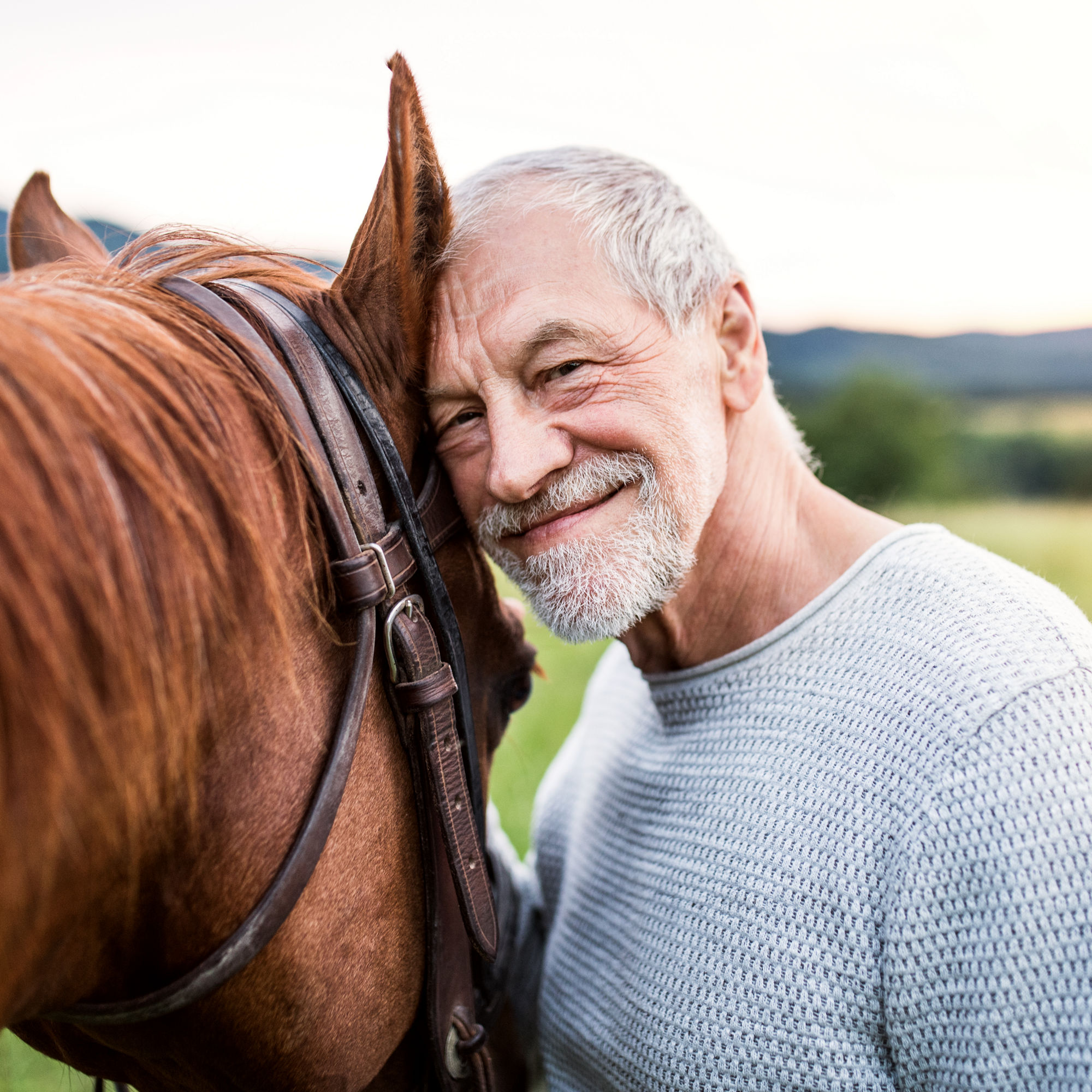 En glad, äldre man lutar sig mot en häst, gröna fält i bakgrunden
