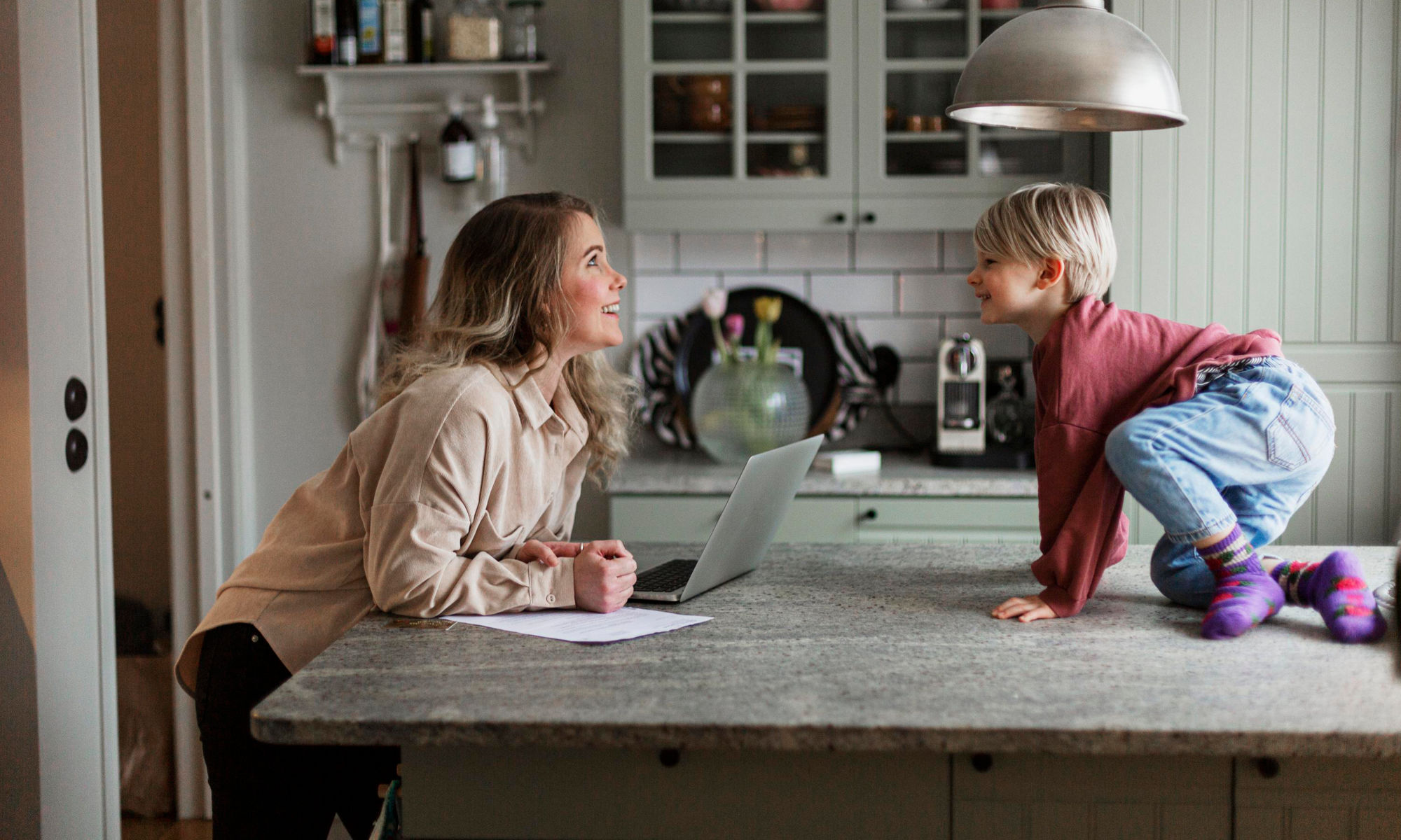 En kvinna sitter vid ett köksbord med en bärbar dator framför sig, och tittar leende på ett barn som sitter på bordet..