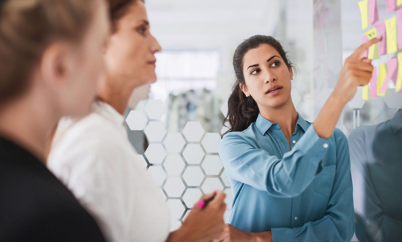 Woman in workshop with two others looking at Post It's on a board.