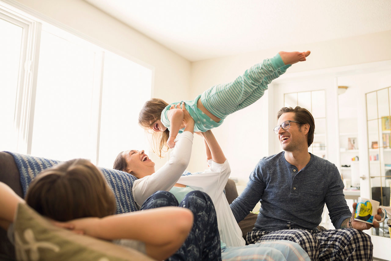 Vuxna och barn sitter i soffan iklädda pyjamas, kvinna lyfter ett barn upp i luften