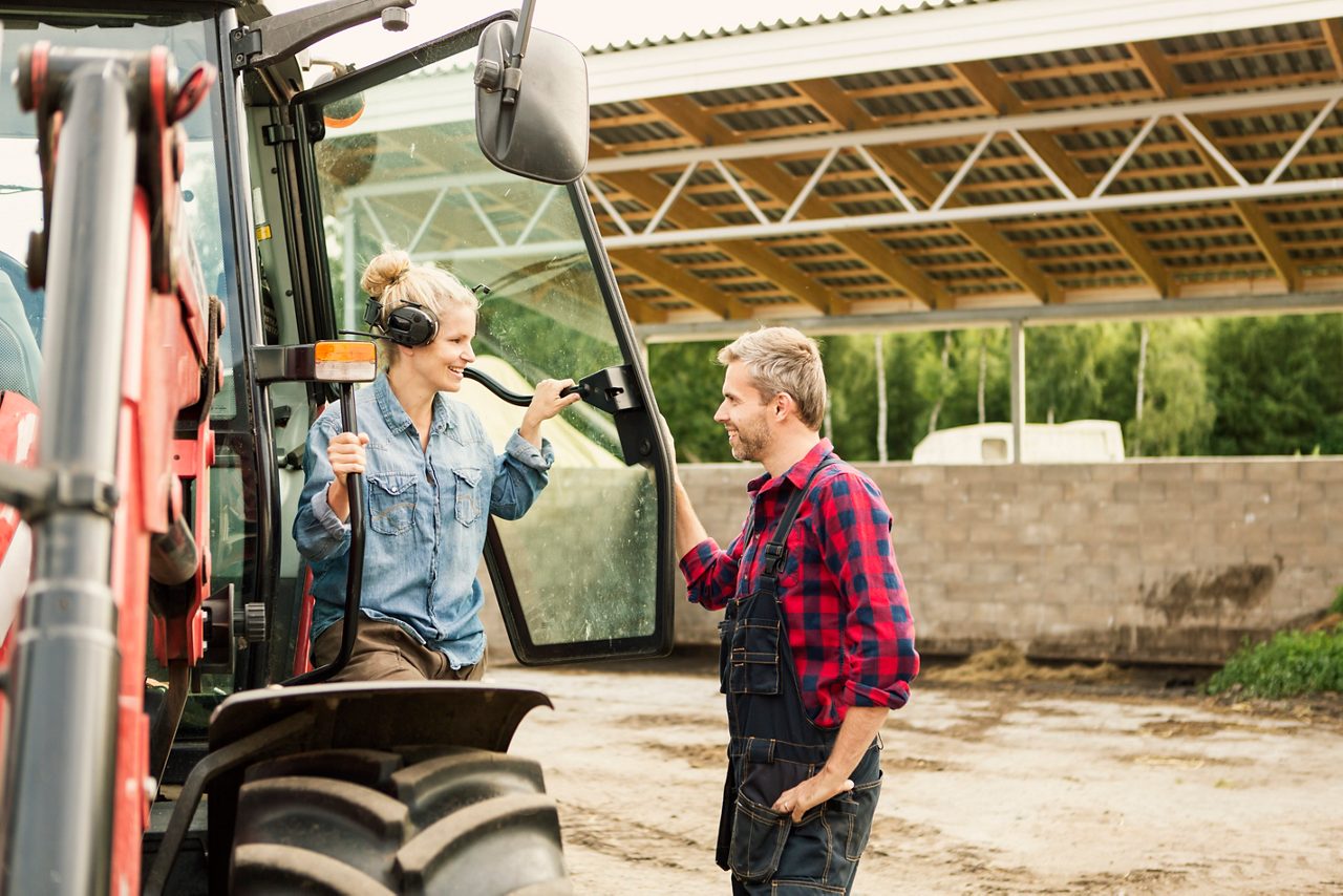 Två bönder tittar leendes på varandra bredvid en traktor
