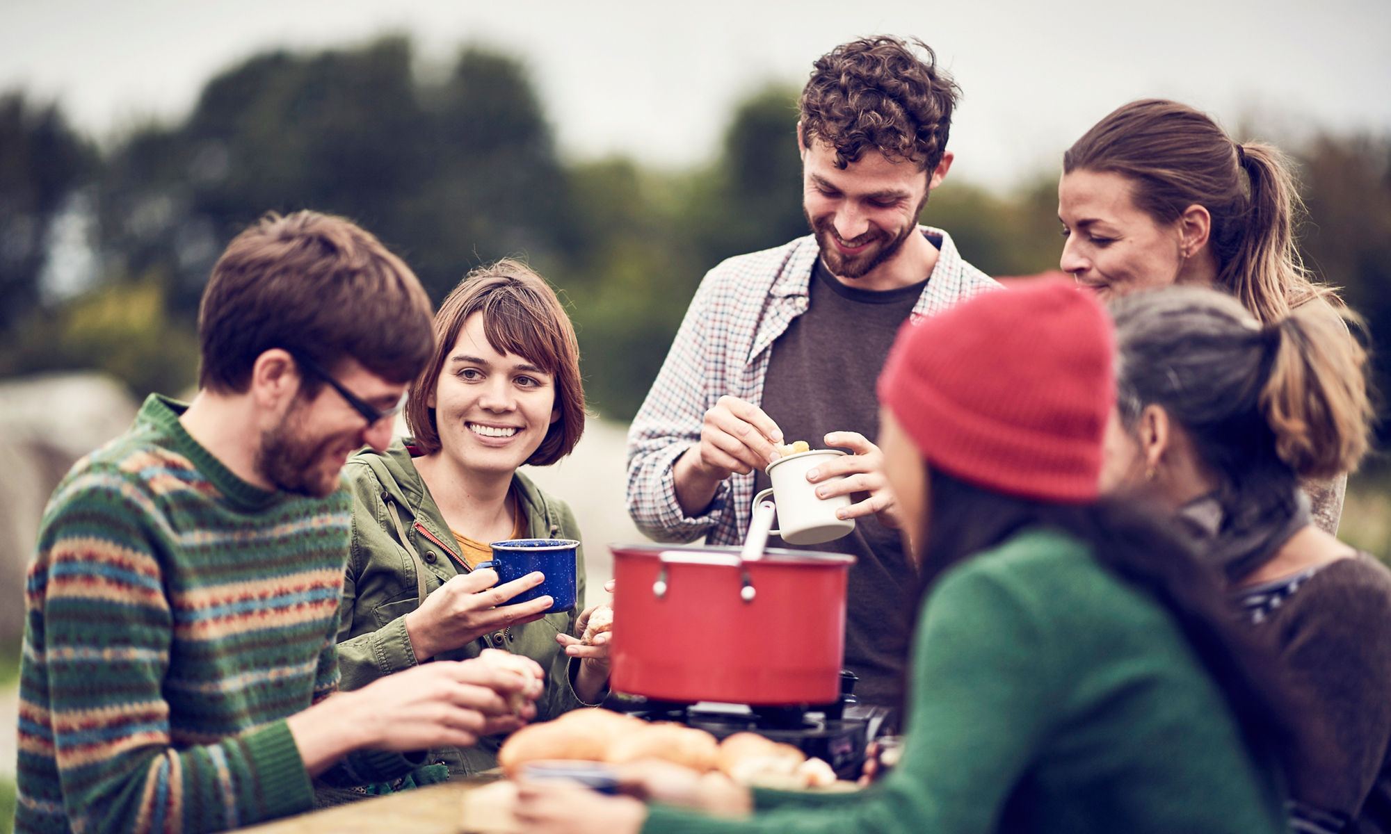 En grupp vuxna sitter utomhus och har picknick, iklädda mössa och stickade tröjor