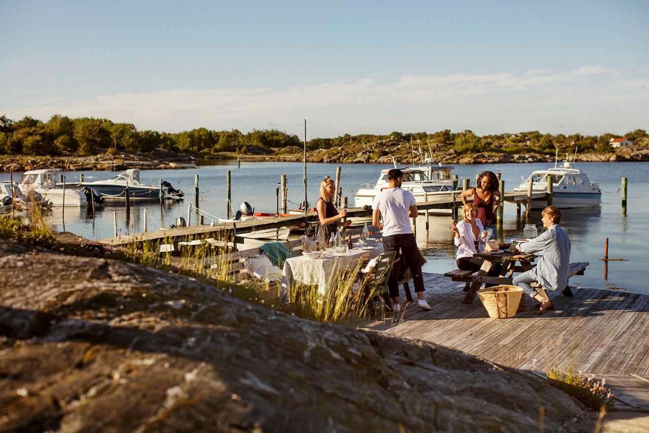 En grupp vuxna sitter vid ett bord ute i en hamn, solen skiner och båtar ligger i vattnet