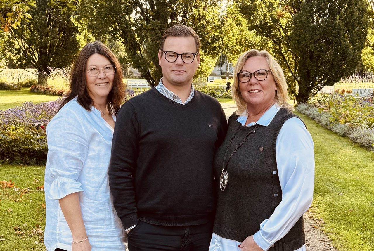Två kvinnor och en man som står i en park och ler mot kameran. 