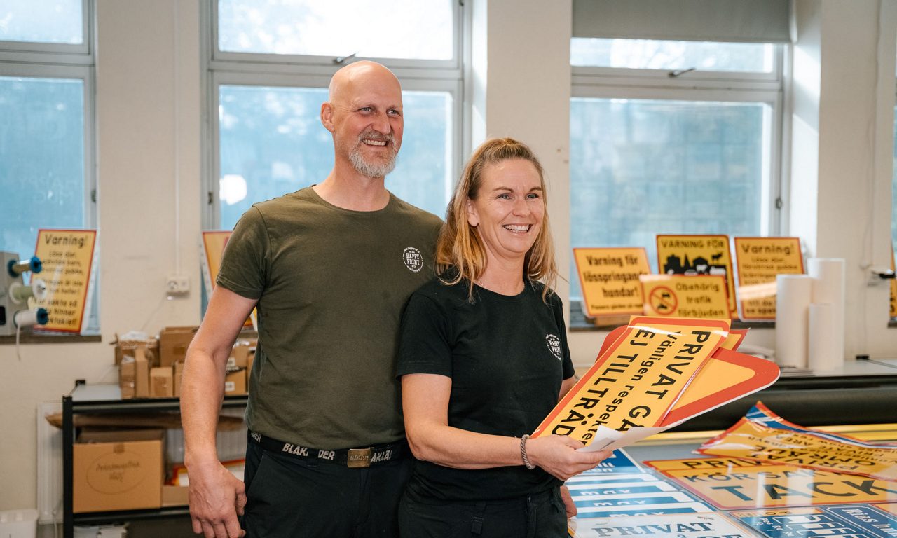 Leende par i tryckeri håller färdigtryckta varningsskyltar.