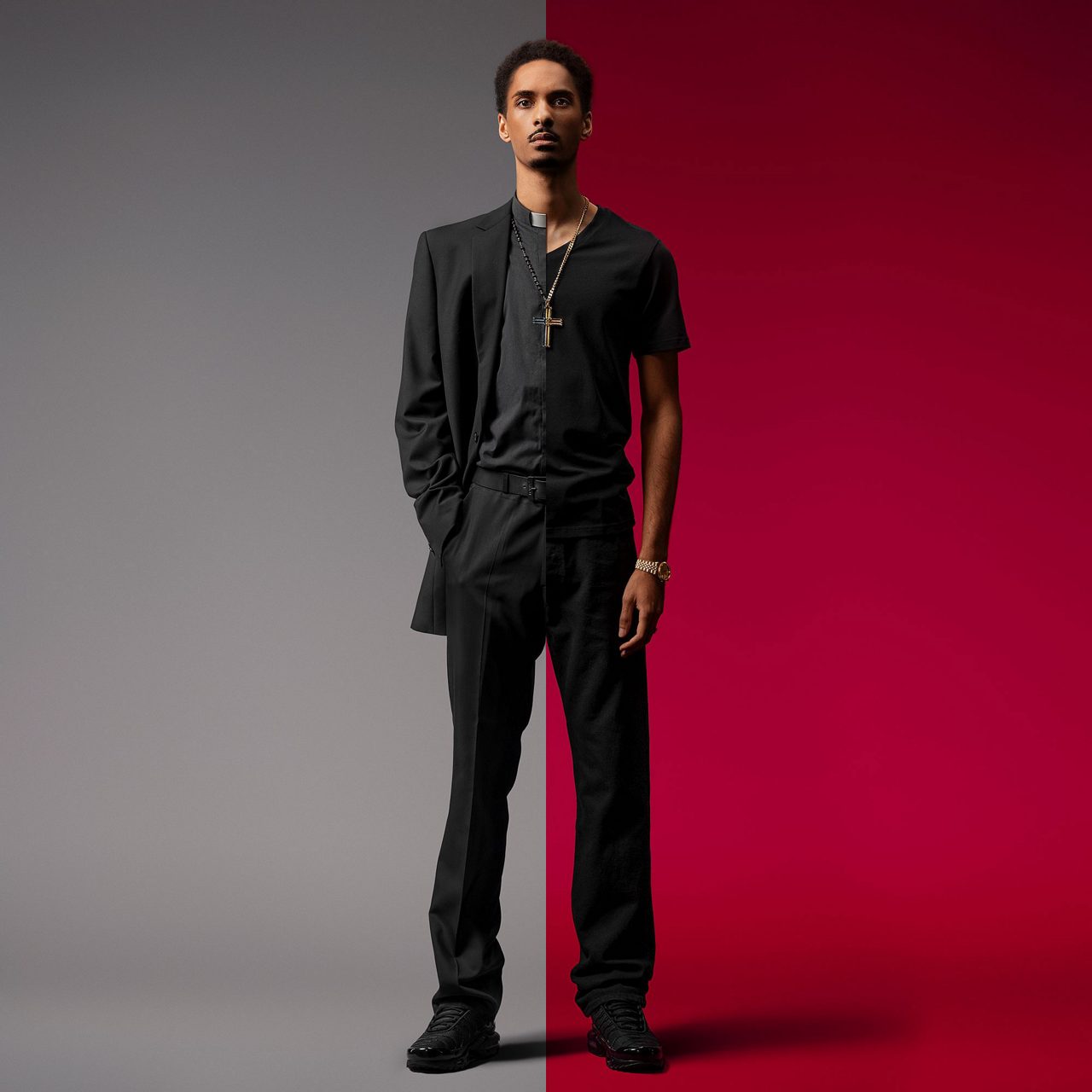 A young man stands confidently wearing a modern black suit and black shoes. He is posed against a smooth gradient backdrop transitioning from gray on the left to deep red on the right. The man wears a cross necklace and a wristwatch, adding subtle metallic accents to his minimalist outfit. The overall scene feels contemporary and fashion-forward, highlighting clean lines and a sleek silhouette.