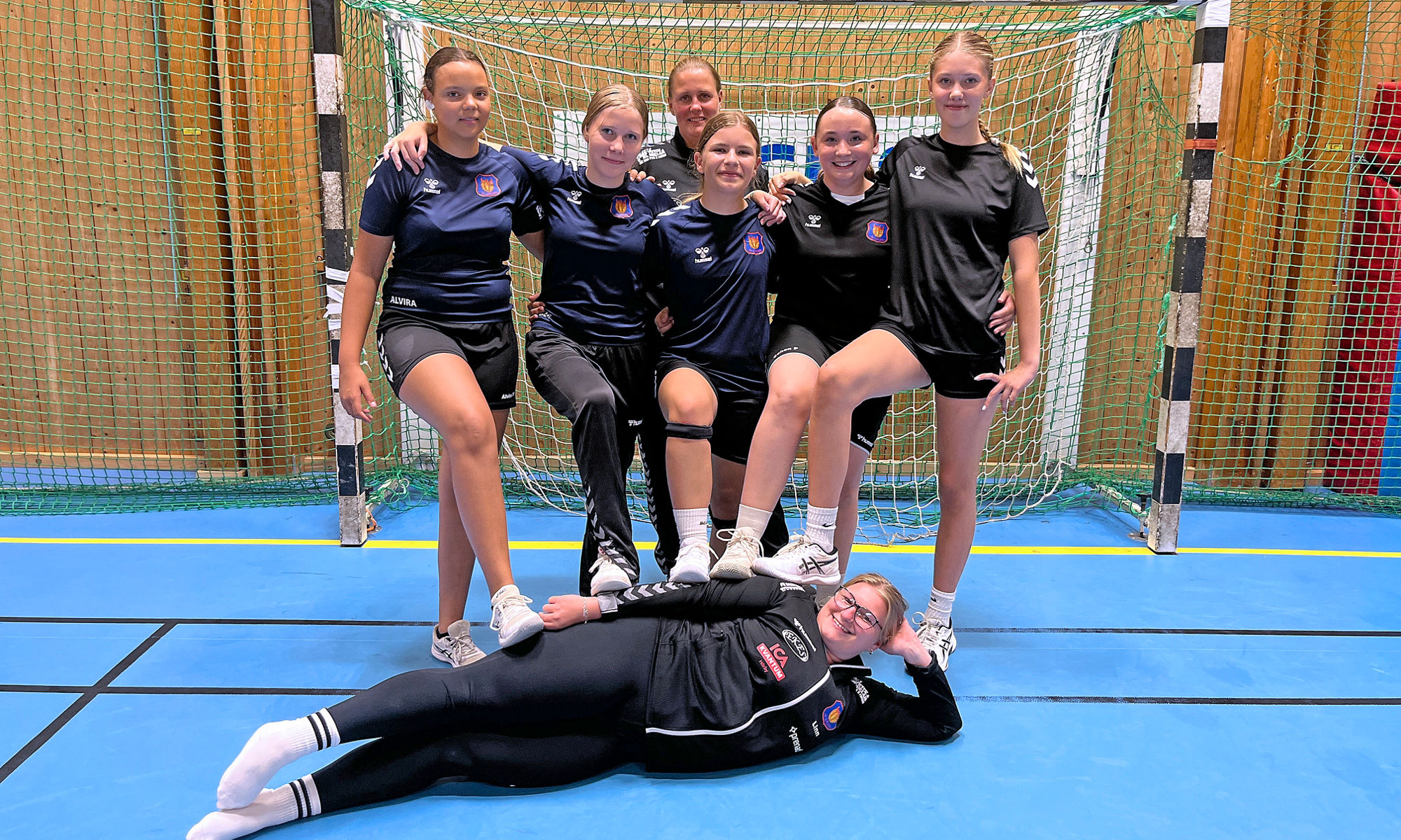 Ett handbollslag med tjejer står i ett handbollsmål och en tjej ligger på golvet.
