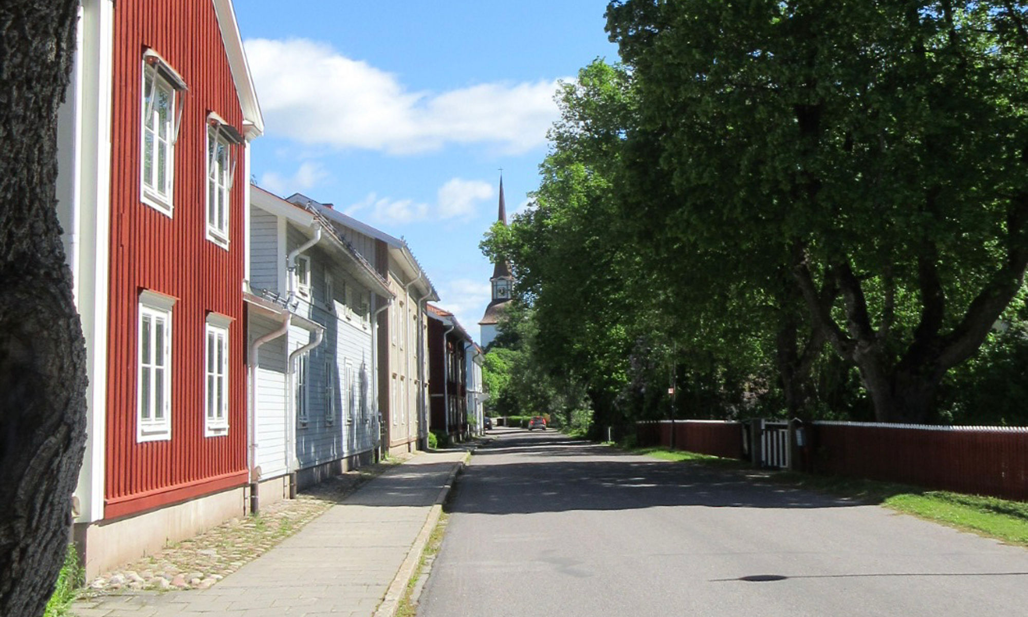 En lugn bostadsgata kantas av traditionella trähus, inklusive en framträdande röd byggnad till vänster. Stora gröna träd välver sig över vägen och skapar ett skuggande tak mot en klarblå himmel. Gatan ser tom och fridfull ut, med en avlägsen kyrkspira synlig i bakgrunden. Solljus framhäver de livfulla färgerna på lövverket och fasaderna, vilket antyder en varm dag i en liten stad eller by.