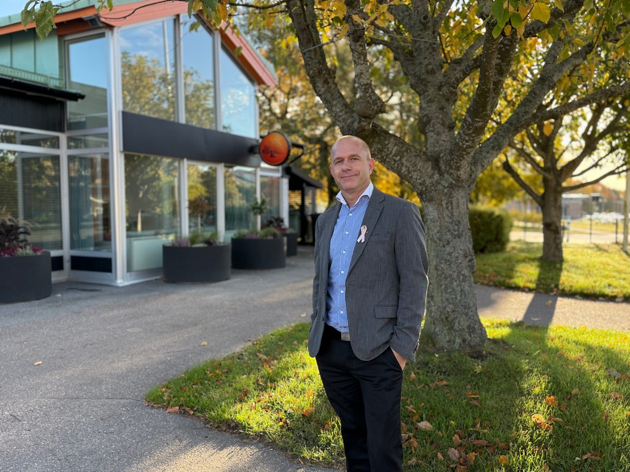 A man in a blue shirt and a grey jacket are standing outside in front of a tree in the morningsun.