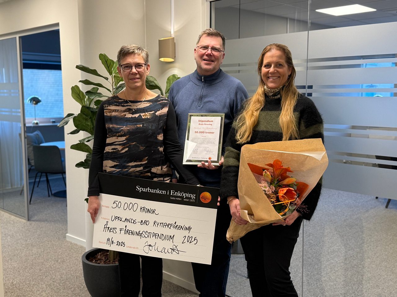 en man och två kvinnor, hållandes stor check, inramat diplom samt blombukett