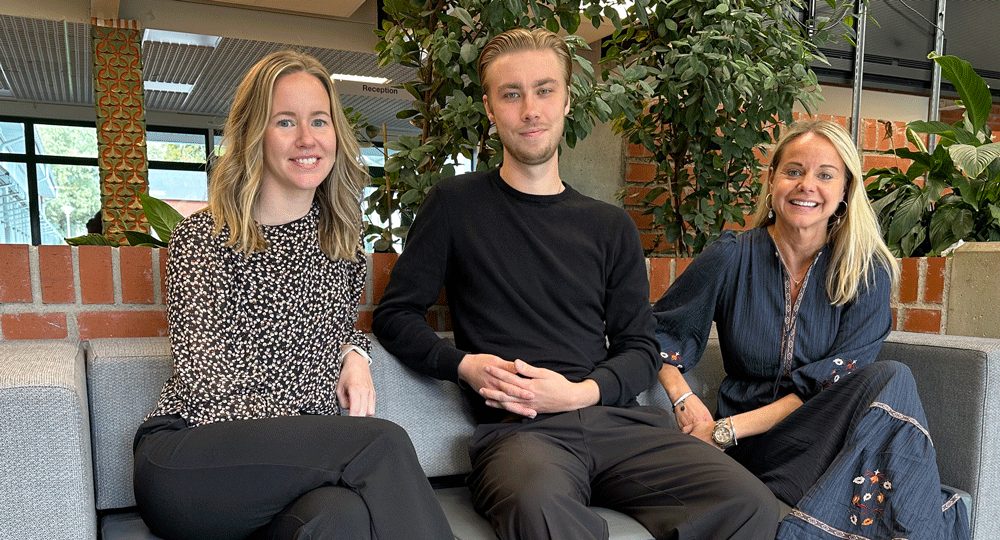 “Tre personer sitter tillsammans i en soffa inomhus, med gröna växter och tegelvägg i bakgrunden.".