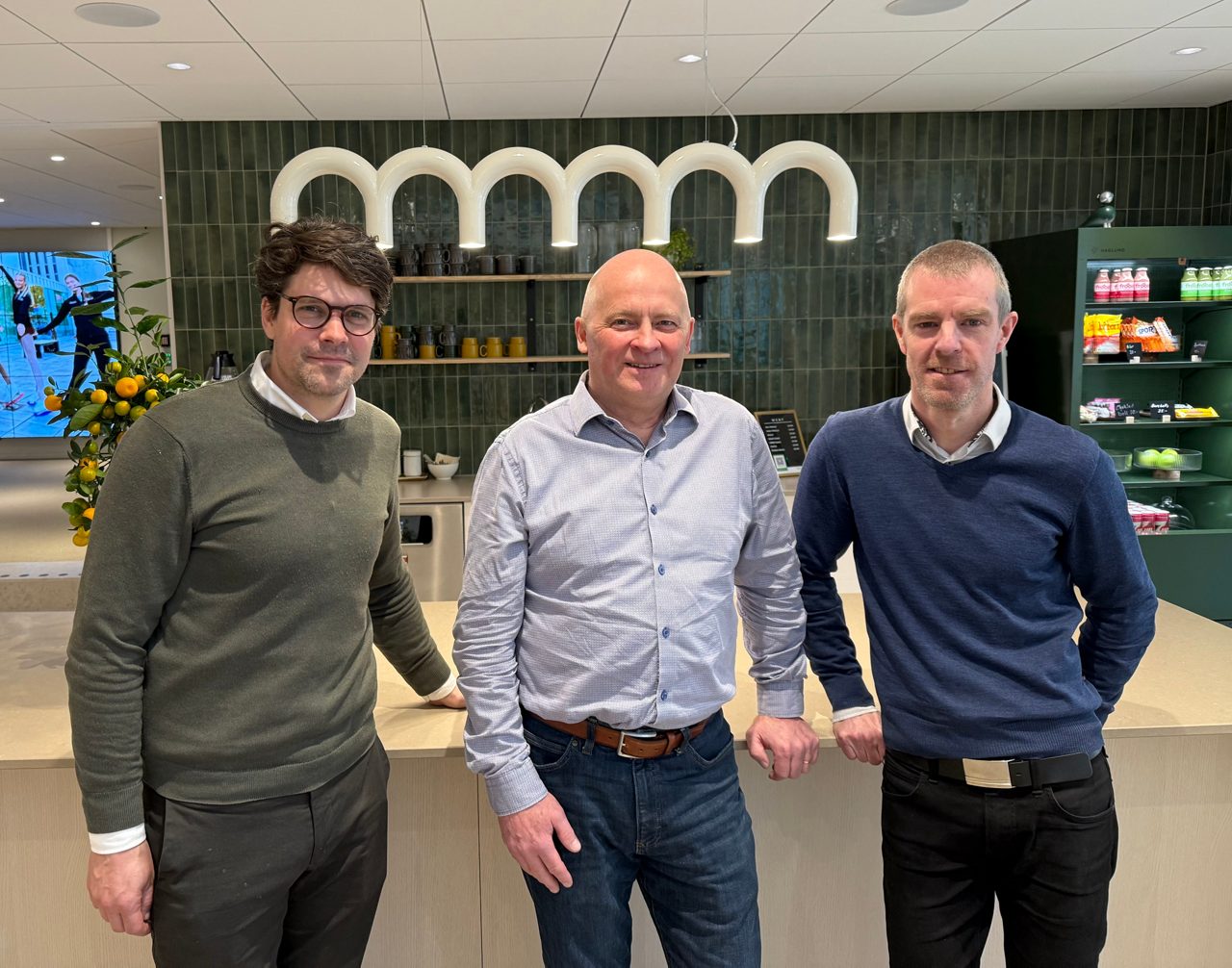 Tre män står vid en köksö i ett cafe, tittar in i kameran och ler