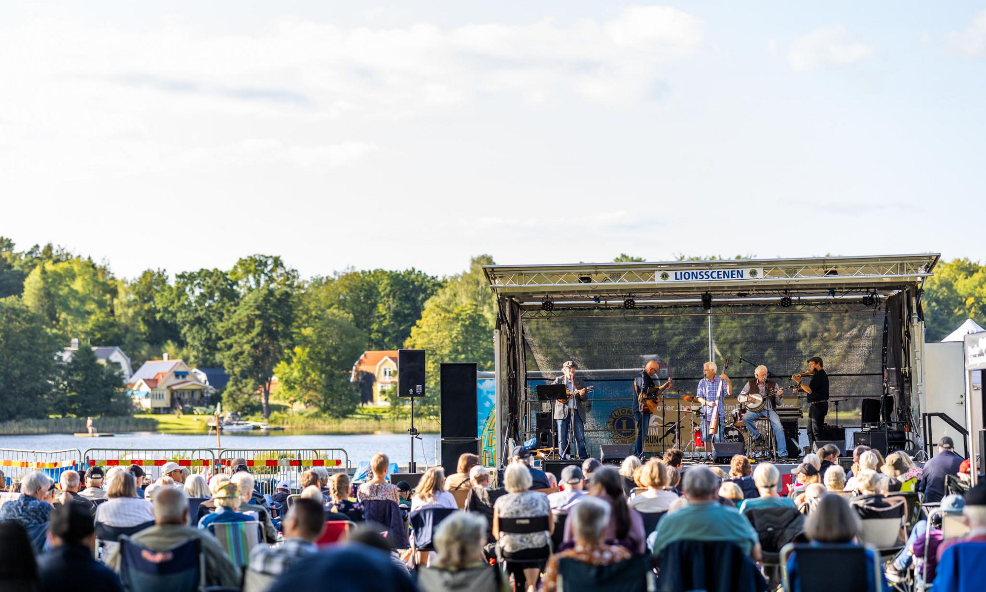 Bilden visar musiker på en scen och en publik som sitter i närheten av en sjö. 