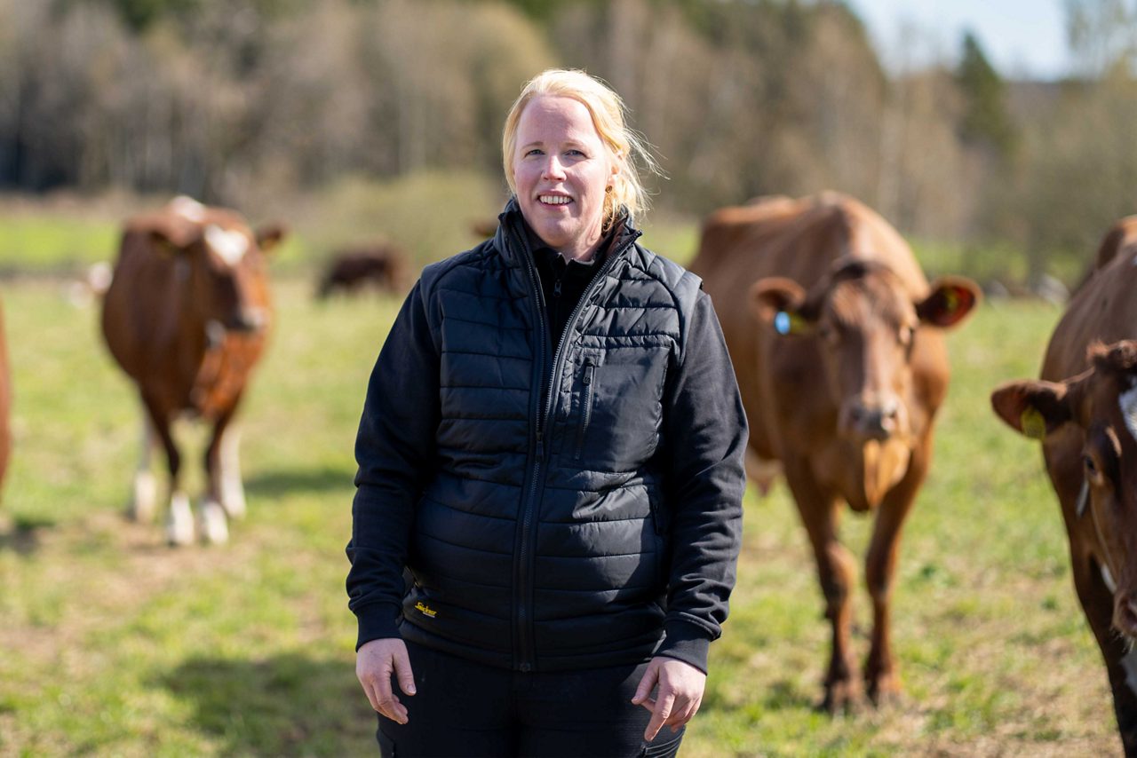 Kvinna står i en hage med kor, ler och tittar in i kameran