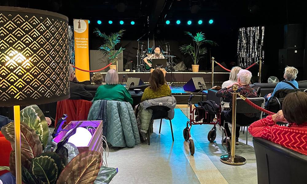 Publikvy från musikcafé med sittande besökare i förgrunden som lyssnar på en artist på scen. På scenen syns mikrofon, instrument och dekor med växter och ljusslingor, och i publiken sitter både äldre och yngre personer, några med rullator. Stämningen är intim och trivsam.