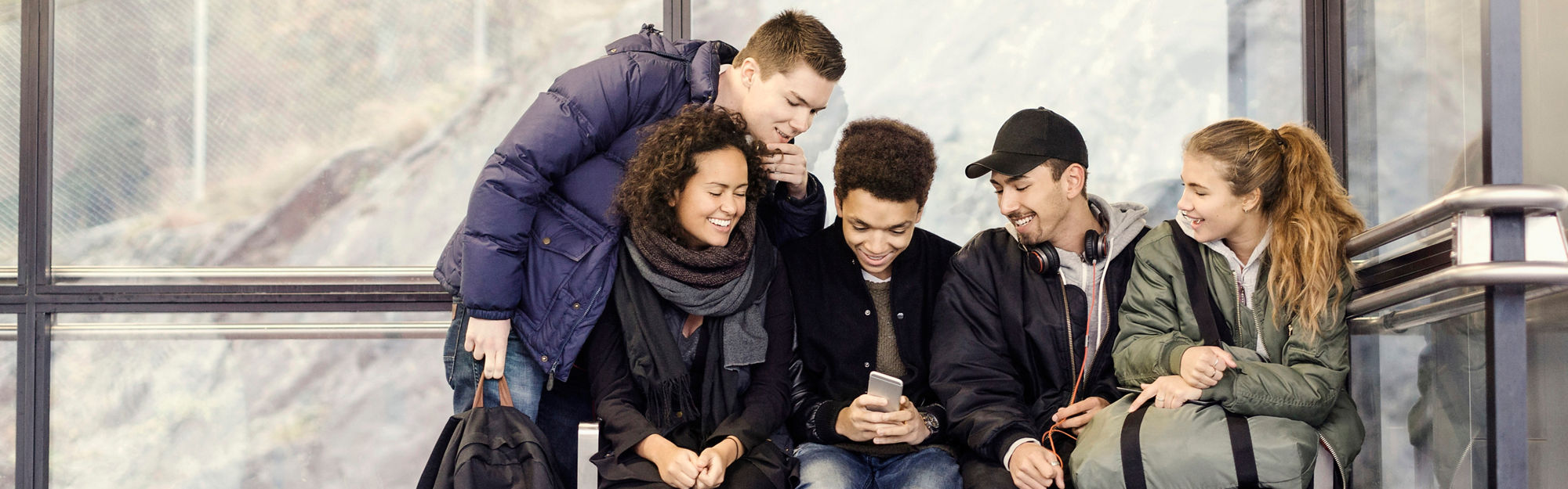 En grupp ungdomar sitter på en busshållplats, ler och tittar på en mobiltelefon
