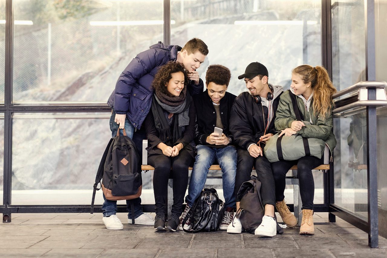 Youths waiting at bus stop in wintertime, smiling while looking at a phone