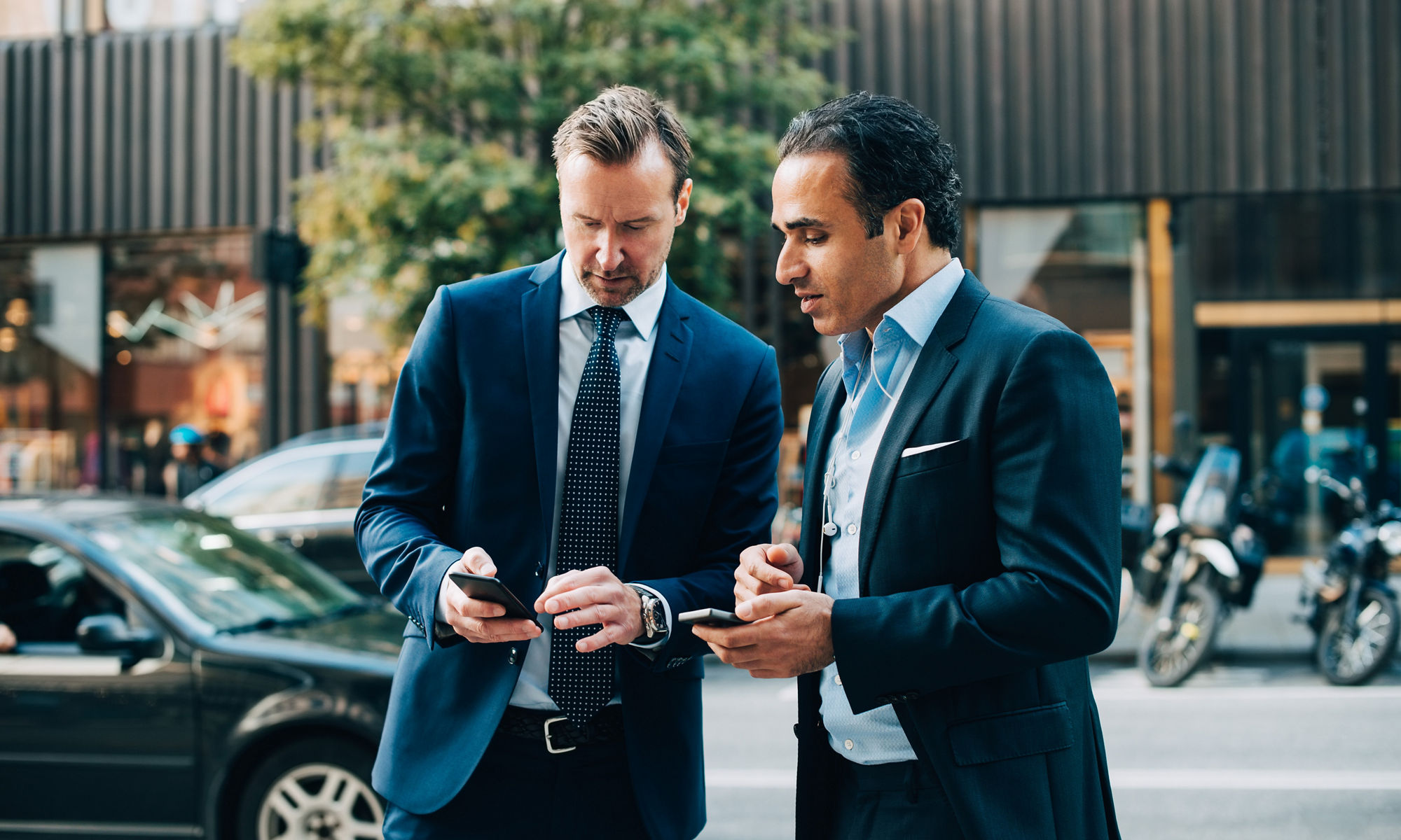 Two men in suits standing outdoors, having a chat while looking at a phone