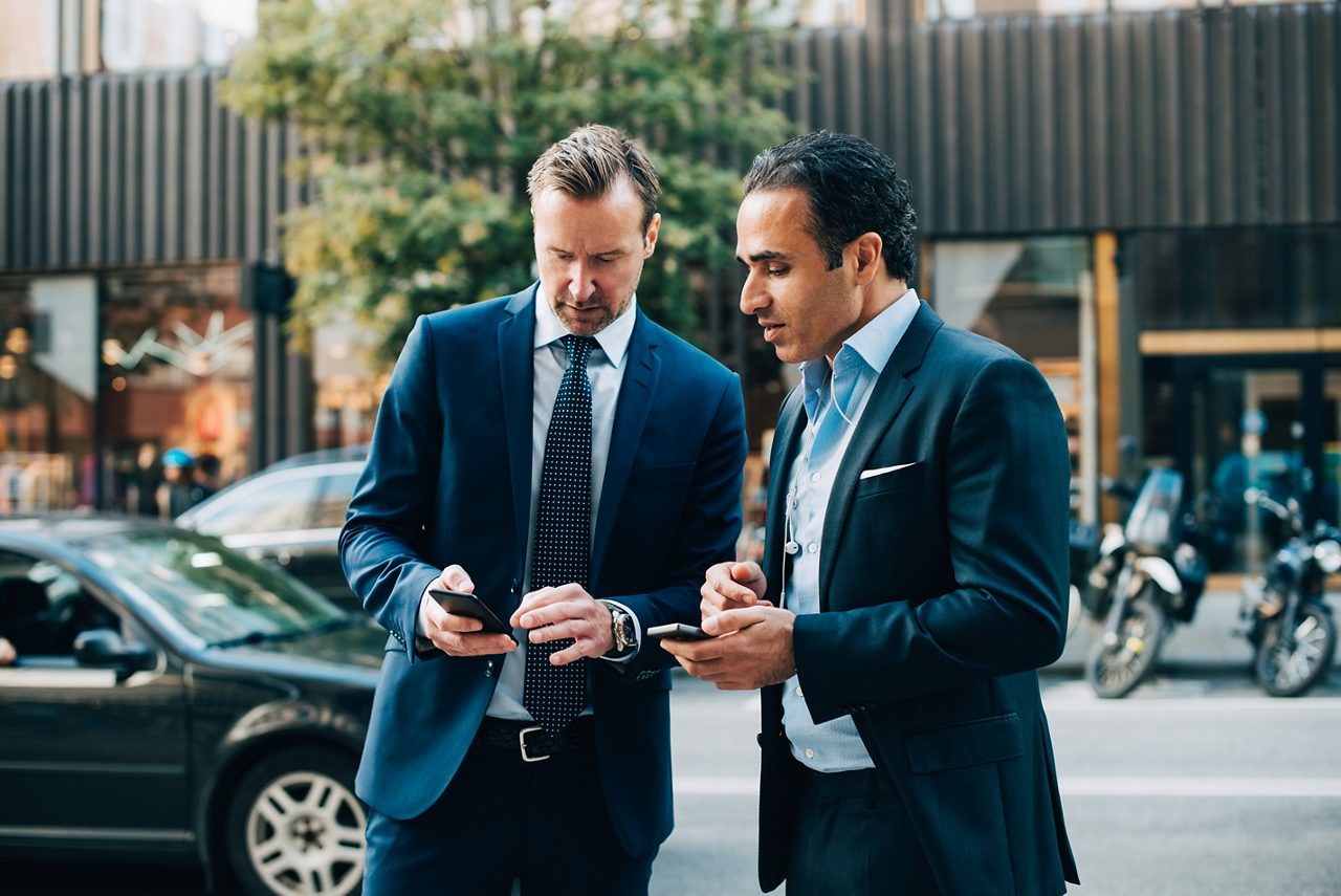 Two men in suits standing outdoors, having a chat while looking at a phone