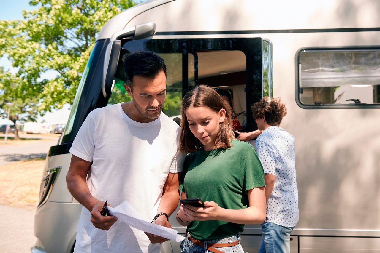 En kvinna och en man utanför en husbil kontrollerar anteckningar på ett papper och en telefon.