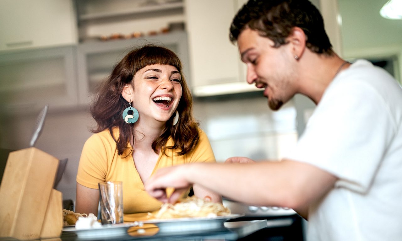 En skrattande tjej och en glad kille sitter vid ett köksbord och äter middag.