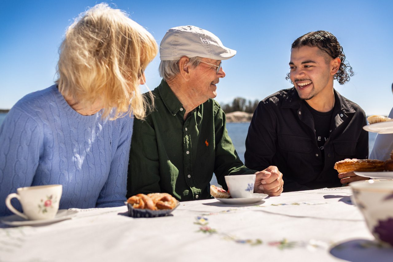 Tre personer sitter utomhus vid ett bord och fikar i solskenet. En äldre man och en kvinna samtalar och ler mot en yngre man, med kaffekoppar och bakverk framför sig.
