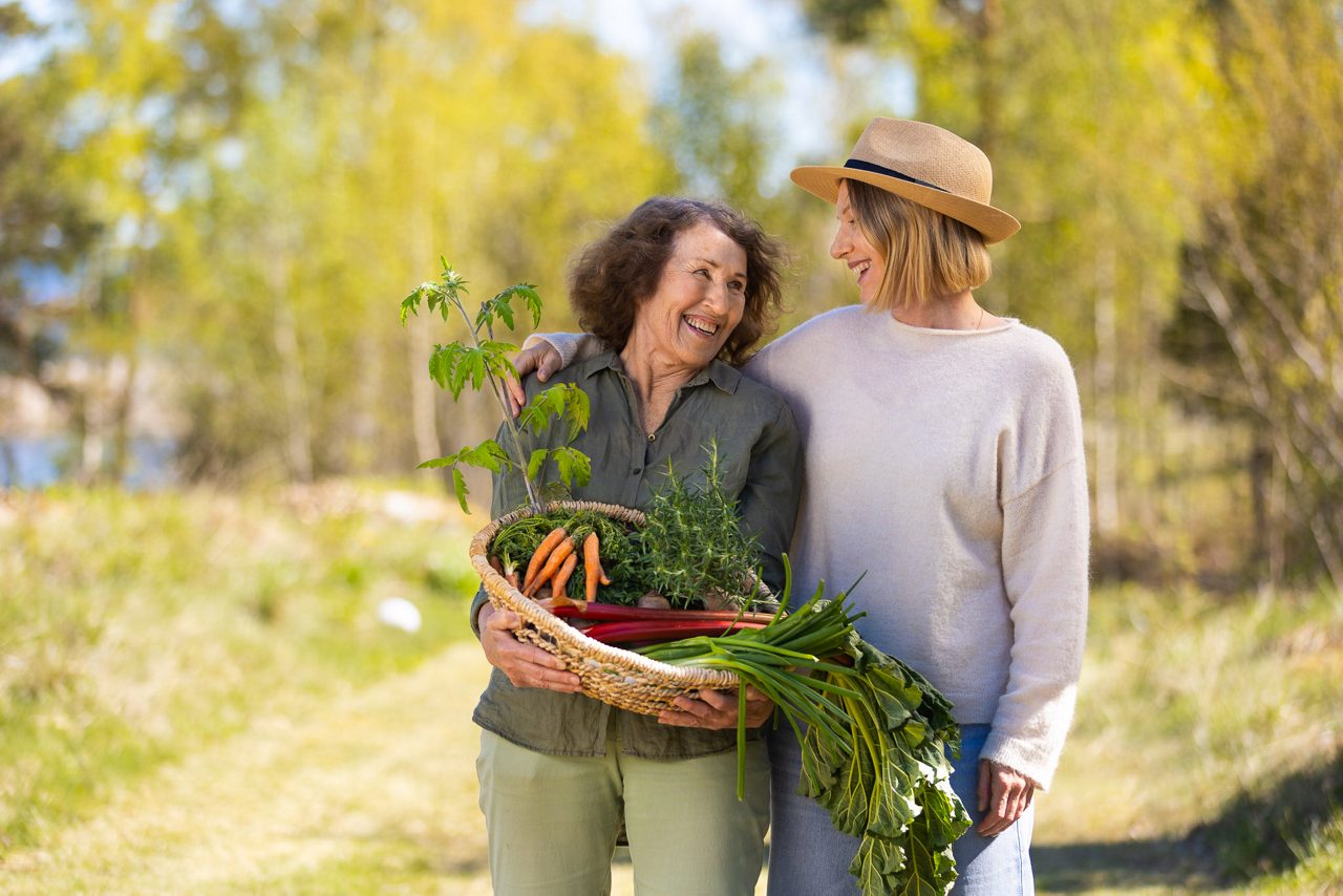 Två kvinnor står utomhus i en solig, grön miljö och ler mot varandra. Den ena håller en korg med nyskördade grönsaker som morötter, örter och tomater, medan de verkar njuta av en stund tillsammans i naturen.